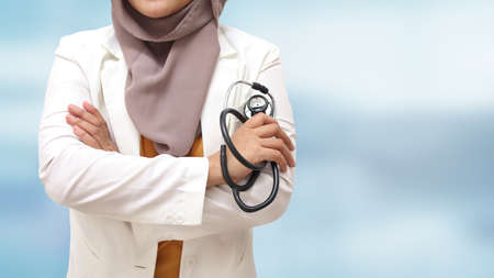 Muslim female doctor wearing hijab and suit, standing with crossed arm and holding stethoscope, succesful confident person, against blue backgroundの写真素材