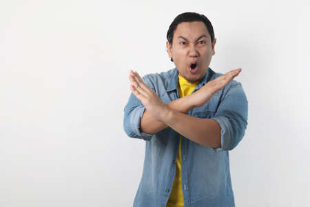 Portrait of young funy angry Asian man shows crossed arms gesture, giving warn to stop gesture,...