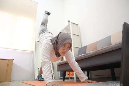 Asian muslim woman wearing hijab doing exercise at home while watching online video instruction on laptop, indoor home workout concept, keep healthy on new normal lifestyleの写真素材