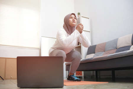 Asian muslim woman wearing hijab doing exercise at home while watching online video instruction on...
