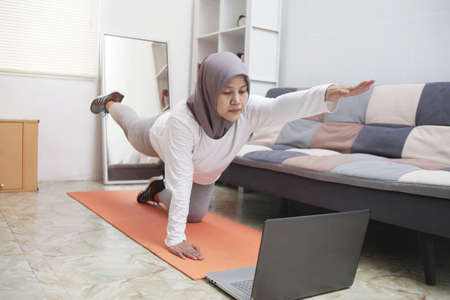 Asian muslim woman wearing hijab doing exercise at home while watching online video instruction on...