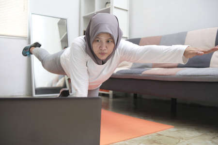 Asian muslim woman wearing hijab doing exercise at home while watching online video instruction on...