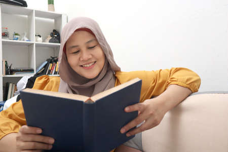 Beautiful Asian muslim woman reading book while sitting on sofa, girl enjoys her time by doing leisure activity, happy cheerful expressionの写真素材