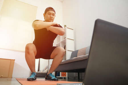 Asian male doing exercise at home while watching online video instruction on laptop, indoor home workout concept, keep healthy on new normal lifestyleの写真素材
