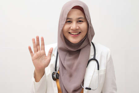 Beautiful young Asian muslim female doctor woman using stethoscope over white isolated background. Waving saying hello happy and smiling, friendly welcome gestureの写真素材
