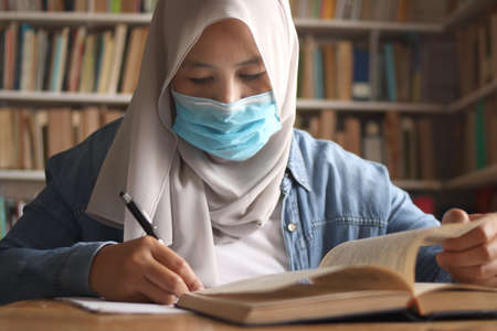 Muslim woman learning studying reading book in library during coronavirus covid19 pandemic, wearing protective medical face maskの写真素材