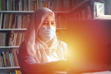 Muslim woman learning studying reading book in library during coronavirus covid19 pandemic, wearing protective medical face maskの写真素材