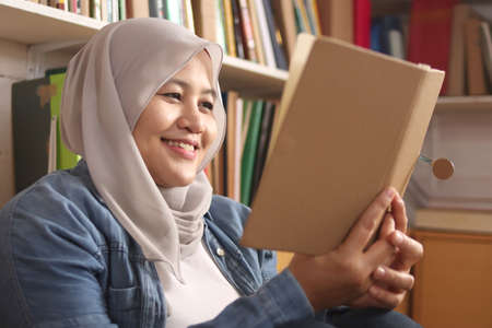 Asian muslim woman wearing hijab reading book in library, educational concept. Happy smiling...