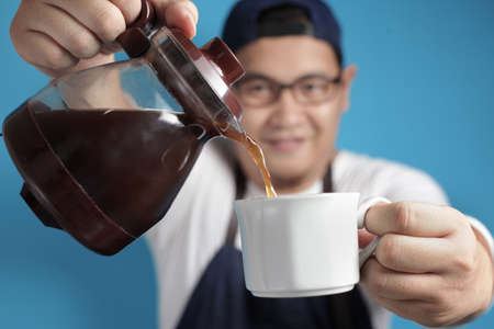 Portrait of Asian male chef or waiter smiling while pouring coffee to a cup, offering coffee concept, against blue backgroundの写真素材