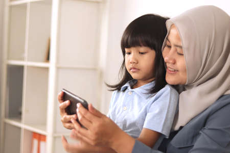 Asian muslim mom and little baby girl daughter learning online or watching videos on tablet phone,...
