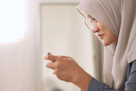 Beautiful Asian muslim woman using phone while relaxing on sofa, playing games or watching streaming...