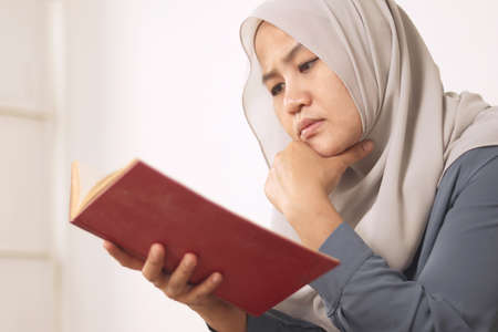 Beautiful Asian muslim woman reading book while sitting on sofa, girl enjoys her time by doing...