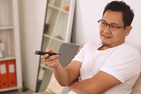 Happy Asian man smiling while watching tv with remote on his hand, sitting at sofa in living roomの写真素材