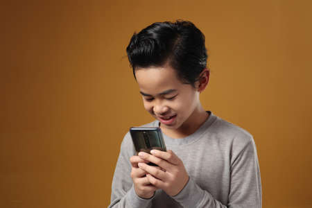 Happy cheerful Asian boy using phone and smiling against yellow background, chat online messaging social media telecommunication conceptの写真素材