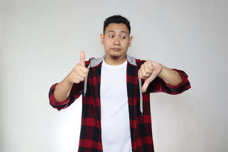 Asian man confused to give bad or good review with thumbs down and up gesture. Making decision conceptの写真素材