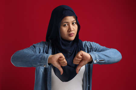 Portrait of muslim lady wearing hijab shows mocking face and thumbs down gesture, disappointed expression, over red backgroundの写真素材
