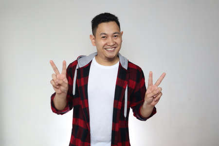 Happy cheerful Asian male smiling and posing at camera, peace sign gesture over grey backgroundの写真素材
