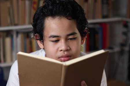 Smart Asian student boy reading a book in library, educational concept, kid learning at homeの写真素材