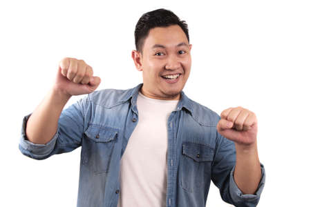 Successful young Asian man shows winning gesture, celebrating victory. Hands raised above with expressive omg wow happy faceの写真素材