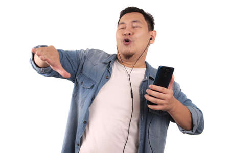 Happy funny young Asian man dancing while listening music on his smart phone with earphones.Isolated on whiteの写真素材