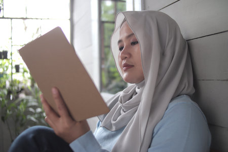 Beautiful Asian muslim woman reading book while sitting outside of her home, girl enjoys her time by doing leisure activity, happy cheerful expressionの写真素材