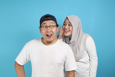 Muslim woman covering her mouth and whispering secret to her husband ear, Asian couple looking shocked and excited, opening eyes and mouth widely with surprise, family conceptの写真素材