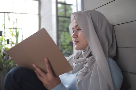 Beautiful Asian muslim woman reading book while sitting outside of her home, girl enjoys her time by doing leisure activity, happy cheerful expressionの写真素材