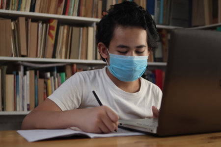 Asian boy student wearing protective mask learning studying online with laptop computer in library, school from home conceptの写真素材