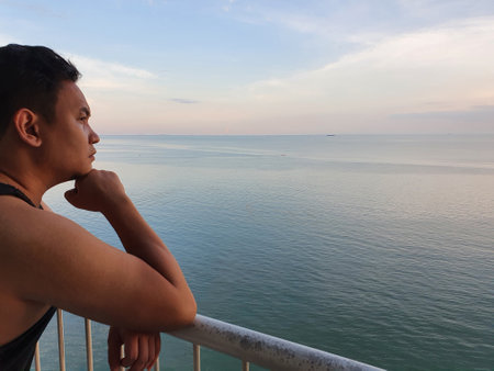 Asian man looking to the ocean from balcony at dawn or sunset, thinking contemplating gestureの写真素材