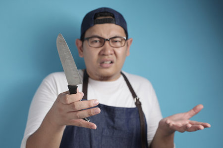 Portrait of Asian male chef confused while holding knife, don't know how to cook, against blue backgroundの写真素材