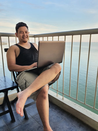 Happy successful Asian man using laptop while sitting on his balcony with calm blue ocean behind, remote working digital nomad conceptの写真素材
