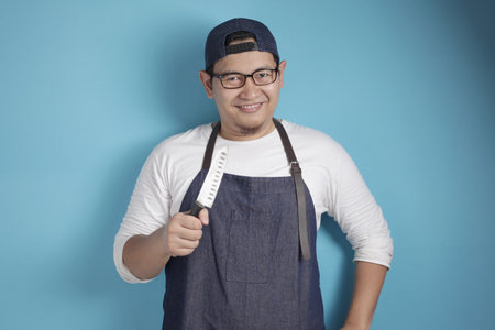 Portrait of Asian male chef smiling while holding knife, ready to cook, against blue backgroundの写真素材