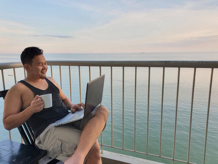 Happy successful Asian man using laptop while sitting on his balcony with calm blue ocean behind, remote working digital nomad conceptの写真素材