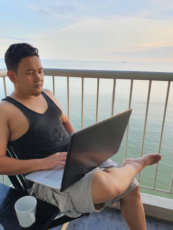 Happy successful Asian man using laptop while sitting on his balcony with calm blue ocean behind, remote working digital nomad conceptの写真素材