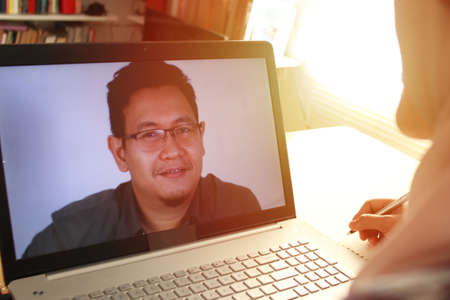 Asian muslim woman having video teleconference witha man on her laptop at home, online learning or...
