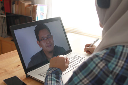Asian muslim woman having video teleconference witha man on her laptop at home, online learning or...