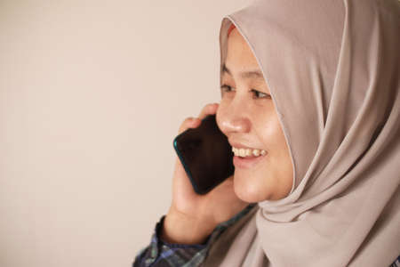 Happy Asian muslim woman wearing hijab talking on phone, good news conversation communication concept, smiling expression, side viewの写真素材