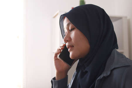 Happy Asian muslim woman wearing hijab talking on phone, good news conversation communication concept, smiling expression, side viewの写真素材