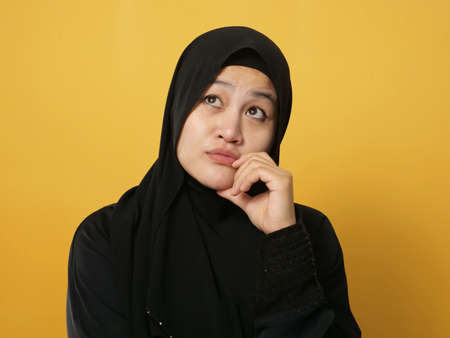 Asian muslim woman wearing hijab shows thinking contemplating expression. Looking for solution of a problem. Close up portrait against yellow backgroundの写真素材