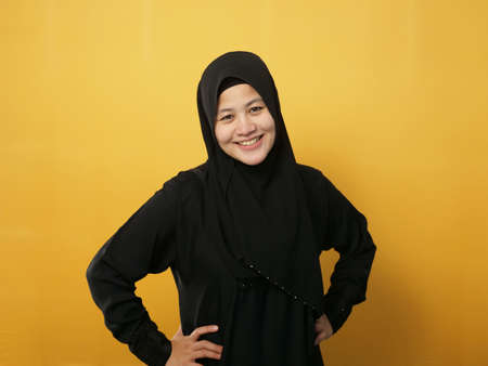 Successful happy Asian muslim woman wearing hijab smiling friendly with hands on hips, confident strong gesture, against yellow backgroundの写真素材