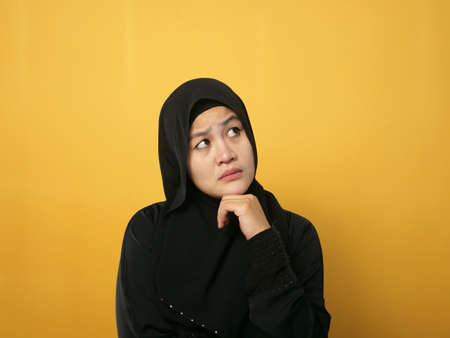 Asian muslim woman wearing hijab shows thinking contemplating expression. Looking for solution of a problem. Close up portrait against yellow backgroundの写真素材