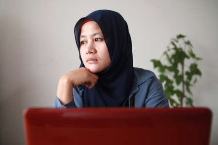 Portrait of Asian muslim woman thinking contemplationg while working on her laptop, sad emotion expressionの写真素材