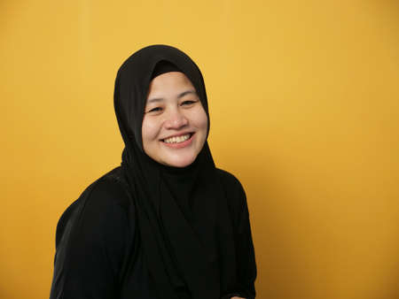 Portrait of happy beautiful young Asian muslim lady wearing hijab smiling and posing for camera, against yellow backgroundの写真素材