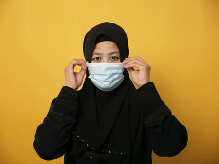 Portrait of Asian muslim woman wearing hijab and facial mask, against yellow backgroundの写真素材