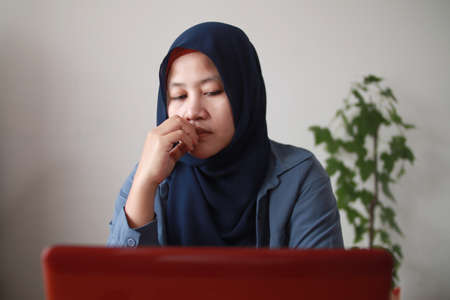 Portrait of Asian muslim woman thinking contemplationg while working on her laptop, sad emotion expressionの写真素材