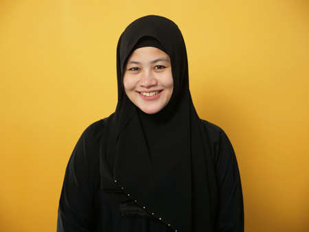 Portrait of beautiful Asian muslim woman wearing hijab smiling at camera, against yellow backgroundの写真素材