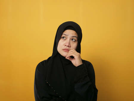 Asian muslim woman wearing hijab shows thinking contemplating expression. Looking for solution of a problem. Close up portrait against yellow backgroundの写真素材