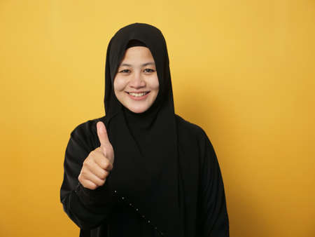 Portrait of young Asian muslim lady wearing hijab shows thumbs up gesture, smiling happy face, approved OK sign, against yellow backgroundの写真素材