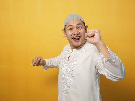 Funny young Asian muslim man dancing happily joyful expressing celebrating good news victory winning success gesture, smiling positive excited emotion, against yellow backgroundの写真素材