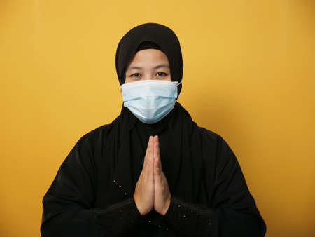 Asian muslim woman wearing hijab and mask during pandemic new normal, woman wearing face protective mask doing greeting gesture, against yellow backgroundの写真素材